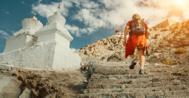 Homem subindo escadas entalhadas em uma montanha. Ao lado dele vemos duas torres brancas, e no topo da montanha, um templo sagrado.