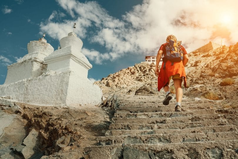 Homem subindo escadas entalhadas em uma montanha. Ao lado dele vemos duas torres brancas, e no topo da montanha, um templo sagrado.