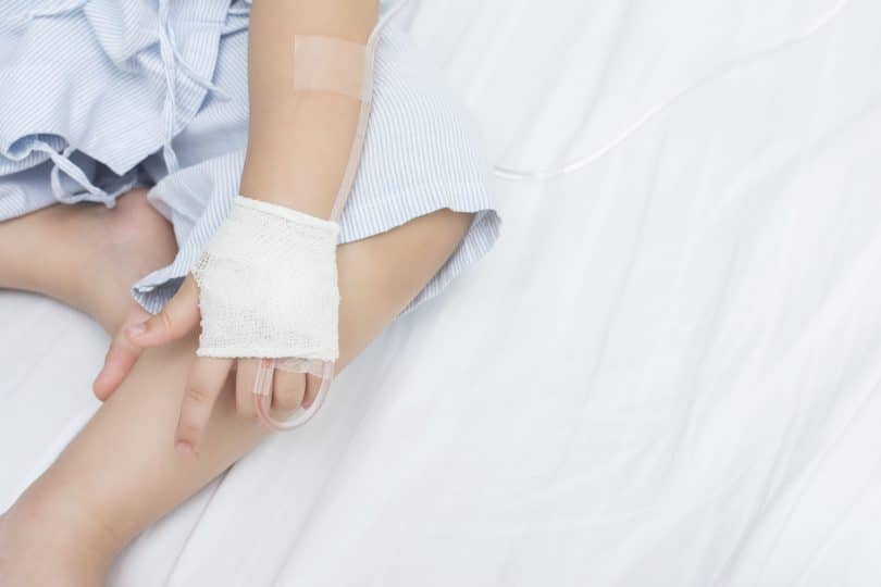 Criança sentada em uma cama, com uma das mãos enfaixada com uma agulha de hemodiálise.