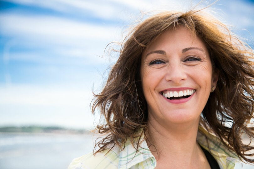 Mulher de meia-idade rindo, com os cabelos ao vento, em frente a um rio em um dia ensolarado.