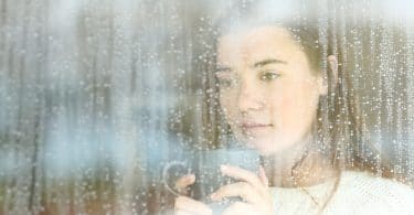 Mulher segurando caneca com as duas mãos e olhando através de janela, molhada do lado de fora pela chuva.