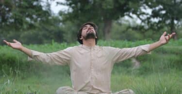 Imagem de um homem meditando em uma área verde. Ele usa roupas em tom de bege claro.