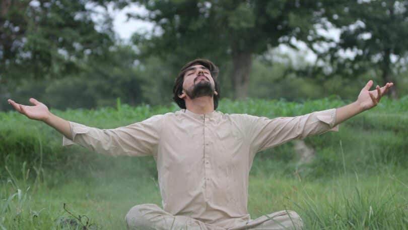 Imagem de um homem meditando em uma área verde. Ele usa roupas em tom de bege claro.