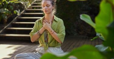 Imagem de uma mulher usando uma roupa na cor bege e uma blusa verde, sentada no chão, fazendo uma meditação em um retiro espiritual.