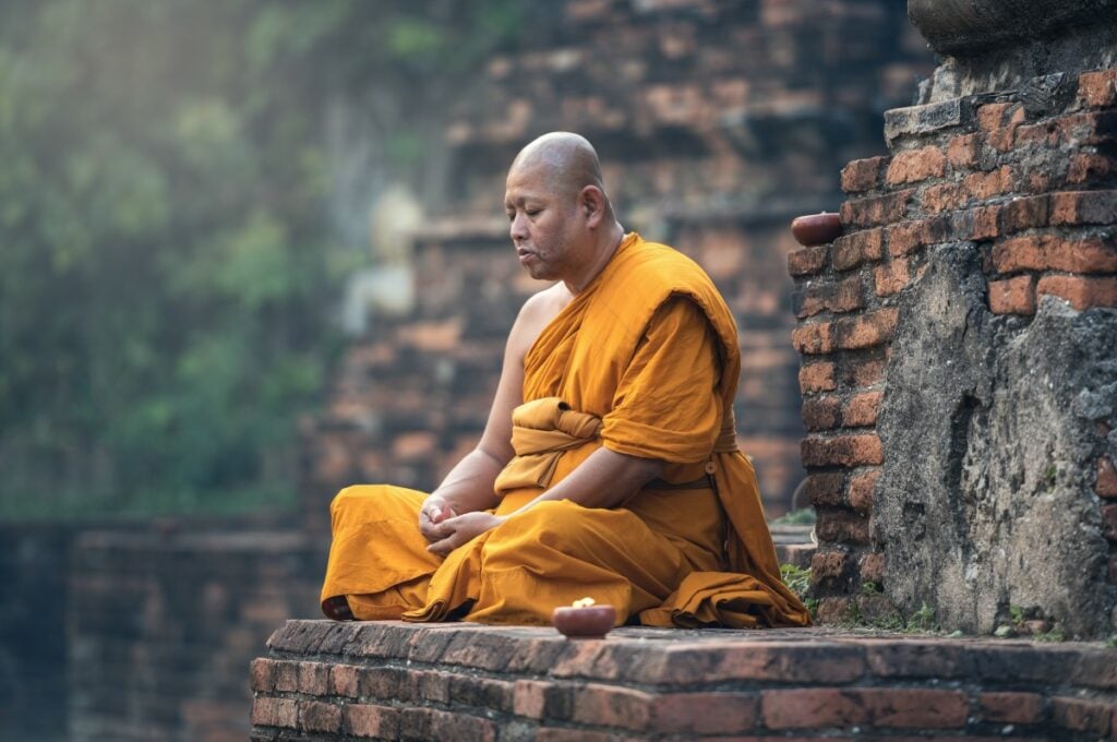 Monge budista meditando em construção antiga. Ele usa trajes típicos laranja, e seus olhos estão fechados.