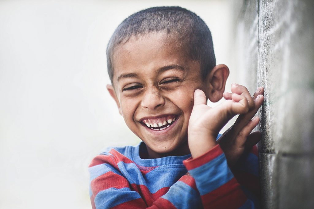 Imagem de uma criança africana vestindo uma blusa de frio listrada nas cores azul claro e vermelho. Ela está muito feliz e sorrindo. Imagem de Pexels por Pixabay.