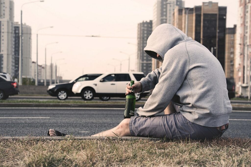 Homem vestindo moletom com capuz e bermuda, sentado na sarjeta, segurando uma garrafa de cerveja.