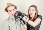 Casal de homem e mulher simulando uma briga. Ela usa uma luva de boxe e está simulando um soco no rosto do homem.