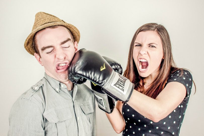 Casal de homem e mulher simulando uma briga. Ela usa uma luva de boxe e está simulando um soco no rosto do homem.