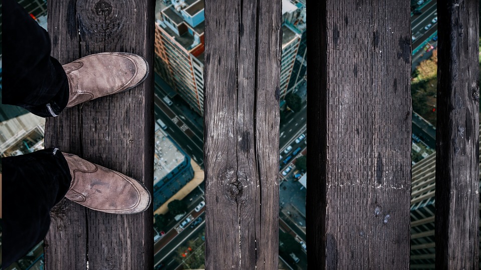 Pés usando sapato marrom em cima de uma placa de madeira. Uma sequência de placas de madeira segue em frente, com um espaço entre elas, como uma ponte. Entre os espaços, é possível ver uma cidade, e um cruzamento de duas avenidas, indicando que a pessoa esteja atravessando em um local muito alto.