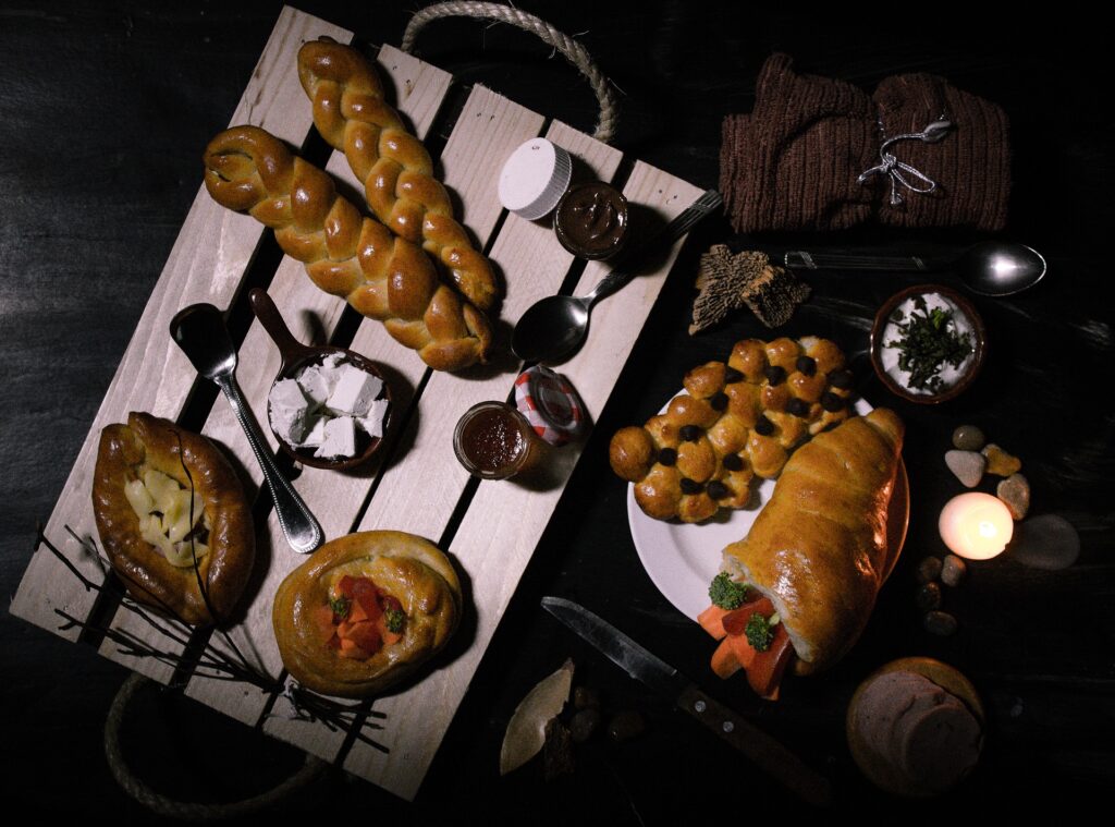 Bandeja de madeira e sobre ela alguns tipos de pães, doces e geleias. Ao lado mais alguns tipos de pães e outros elementos decorativos. 