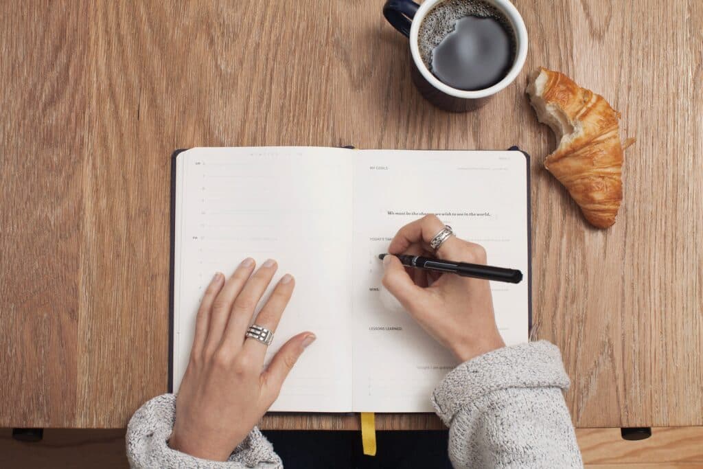 Mulher escrevendo em um caderno sobre uma mesa de madeira, ao lado de uma xícara de café e um croissant.