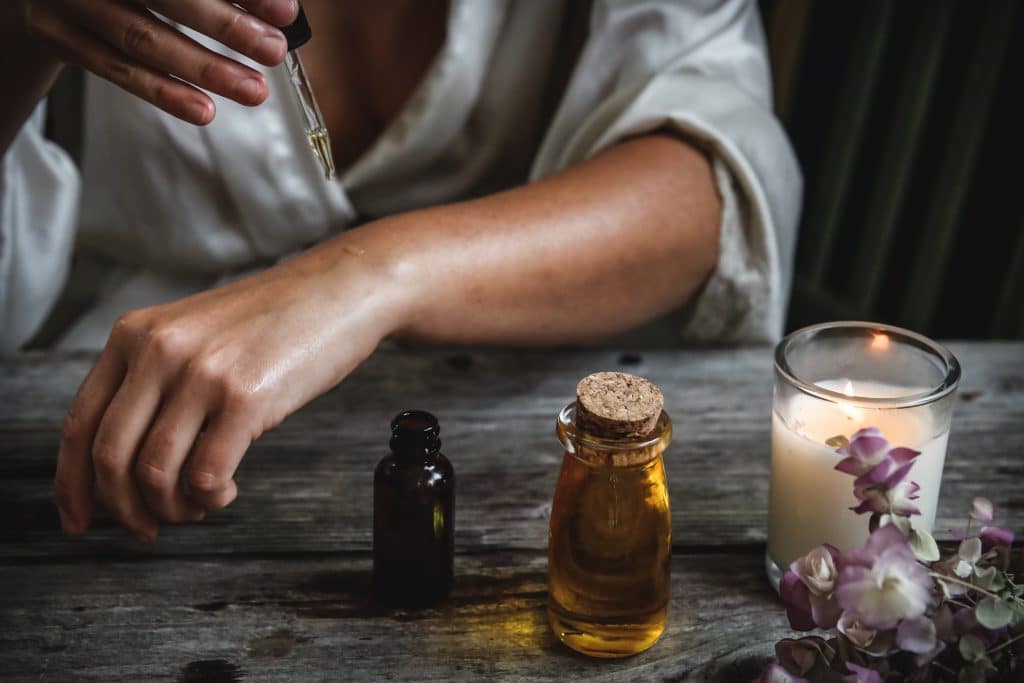 Pessoa aplicando gotas de óleo com um conta-gotas em seu braço. Ao lado, há uma mesa de madeira com frascos de óleo, uma vela acesa e algumas flores.