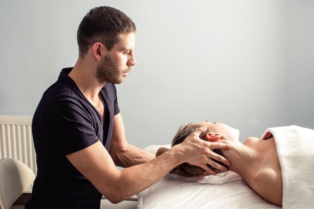 Homem terapeuta segurando a cabeça de uma paciente mulher, que está deitada e com uma toalha sobre o corpo.