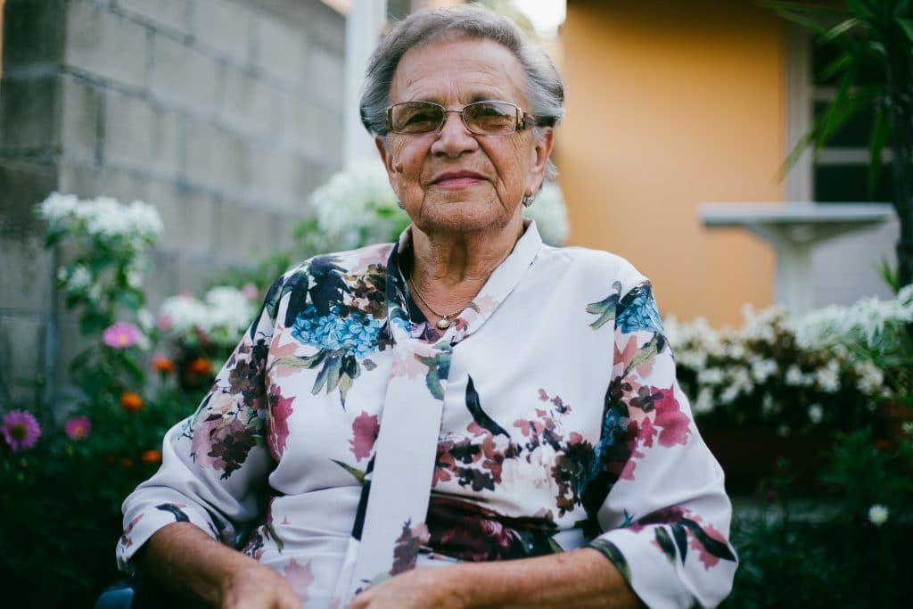 Mulher idosa sentada em um quintal com flores, de óculos, com um leve sorriso no rosto.