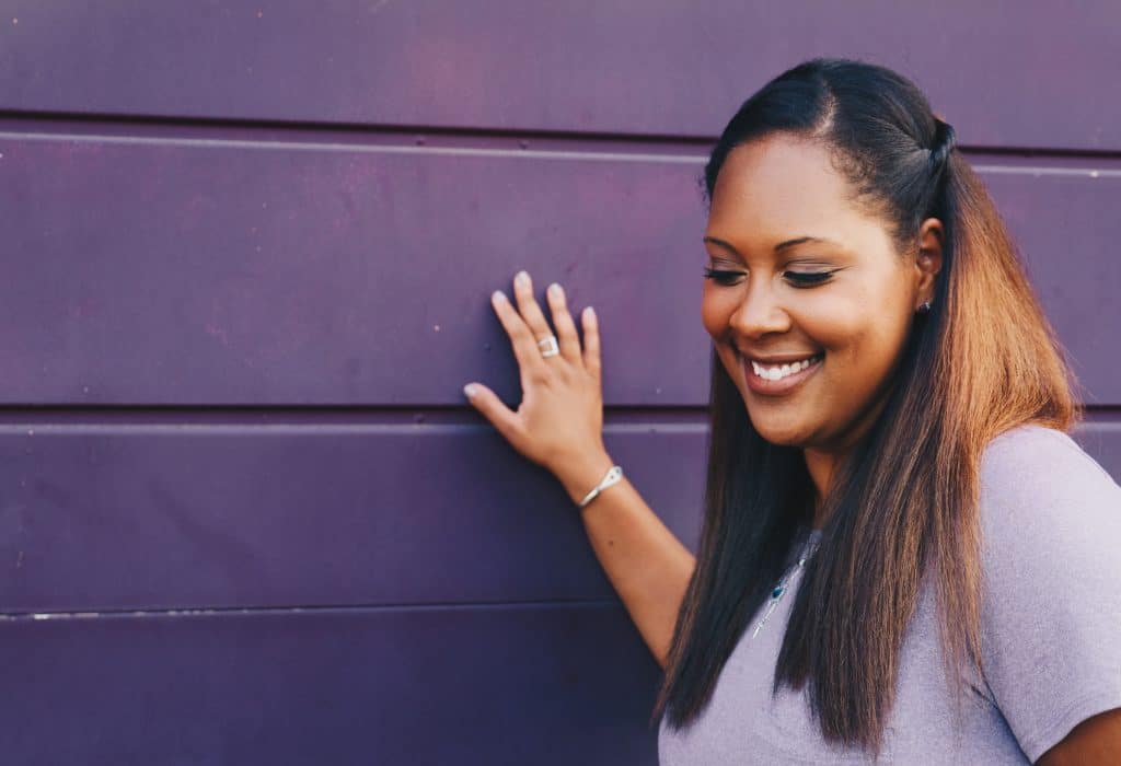 Mulher adulta sorrindo enquanto olha para baixo e apoia a mão sobre uma parede.