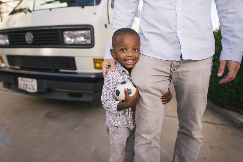 Criança segurando bola de futebol e abraçando perna do pai.