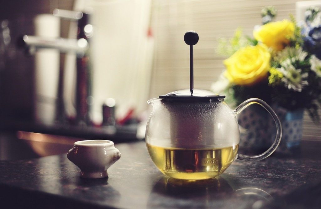 Jarra com chá sobre uma mesa de pedra, ao lado de um pequeno recipiente e de flores em um vaso.