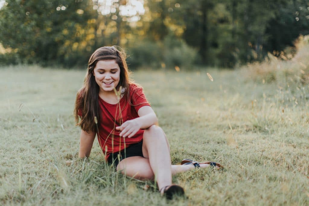 Menina adolescente sentada em um campo, de pernas cruzadas, rindo enquanto olha para baixo.