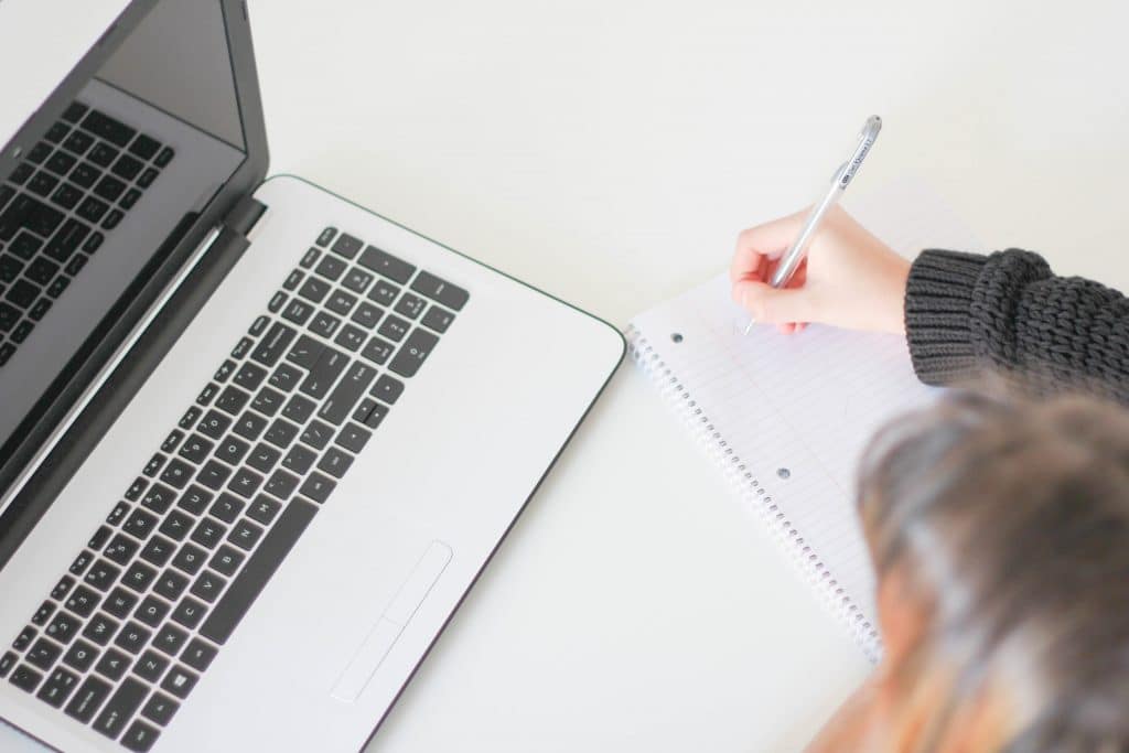 Menina escrevendo em um caderno sobre uma mesa com um computador.