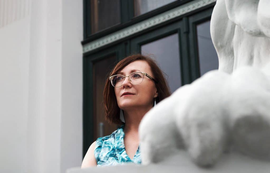 Mulher de meia idade e cabelos lisos, de óculos, olhando para cima em frente a uma construção urbana.