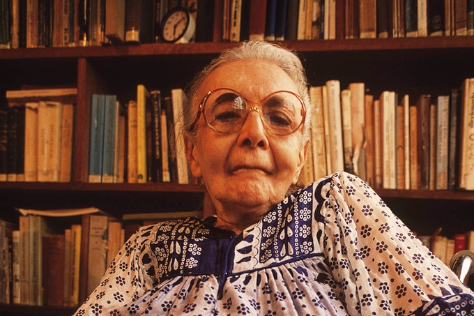 Fotografia de Nise da Silveira (1905-1999) em frente a uma estante de livros.