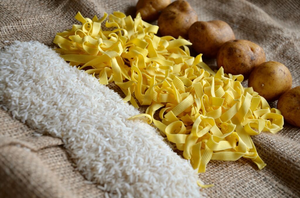 Arroz branco, massa de macarrãp e batatas enfileiradas, lado a lado, dispostas sobre uma mesa coberta com pano de juta. Todos estão crús.
