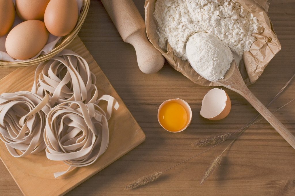 Macarrão tipo Fetuchine. Ao lado uma cesta de ovos vermelhos inteiros. Um rolo de macarrão de madeira, um pacote de farinha de trigo branca, ramos de trigo, um ovo quebrado.