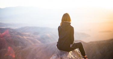 Mulher de costas para a câmera, sentada em uma pedra no topo de uma montanha. Ela veste jaqueta, calça e tênis. Ao fundo, muitas montanhas menores, o mar e o brilho do sol que dificulta a visão do lado direito superior da imagem.