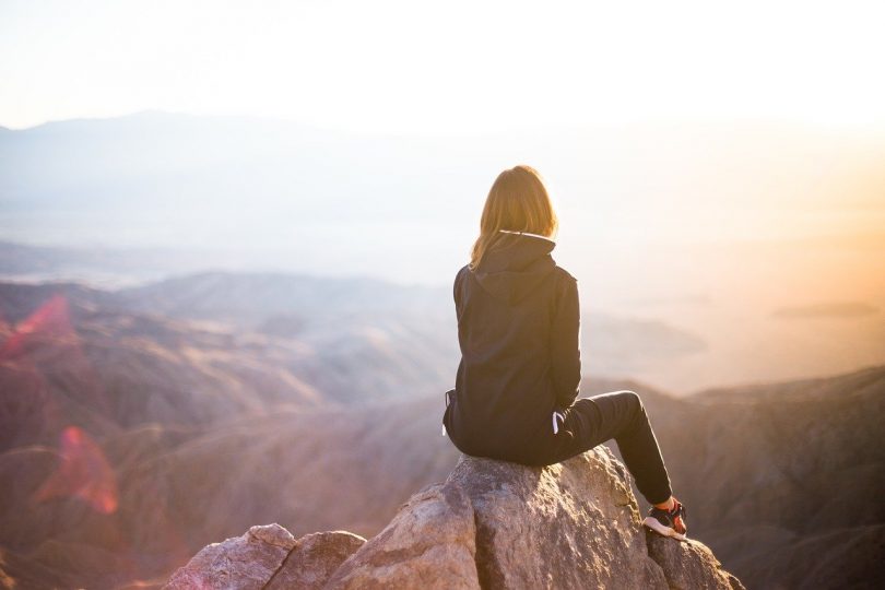 Mulher de costas para a câmera, sentada em uma pedra no topo de uma montanha. Ela veste jaqueta, calça e tênis. Ao fundo, muitas montanhas menores, o mar e o brilho do sol que dificulta a visão do lado direito superior da imagem.