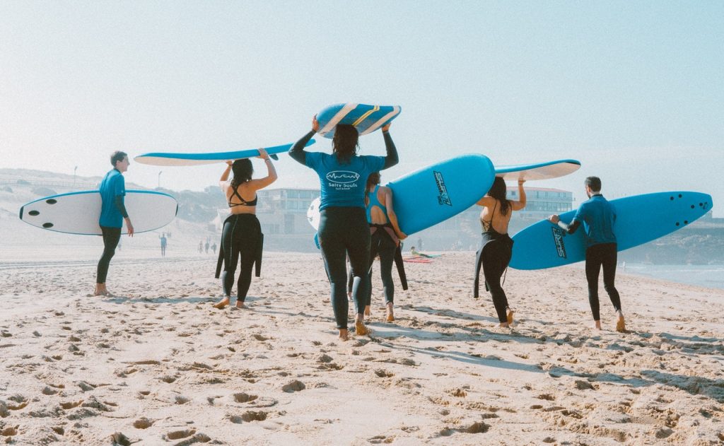 Seis pessoas segurando uma prancha de surfe enquanto caminham na praia.