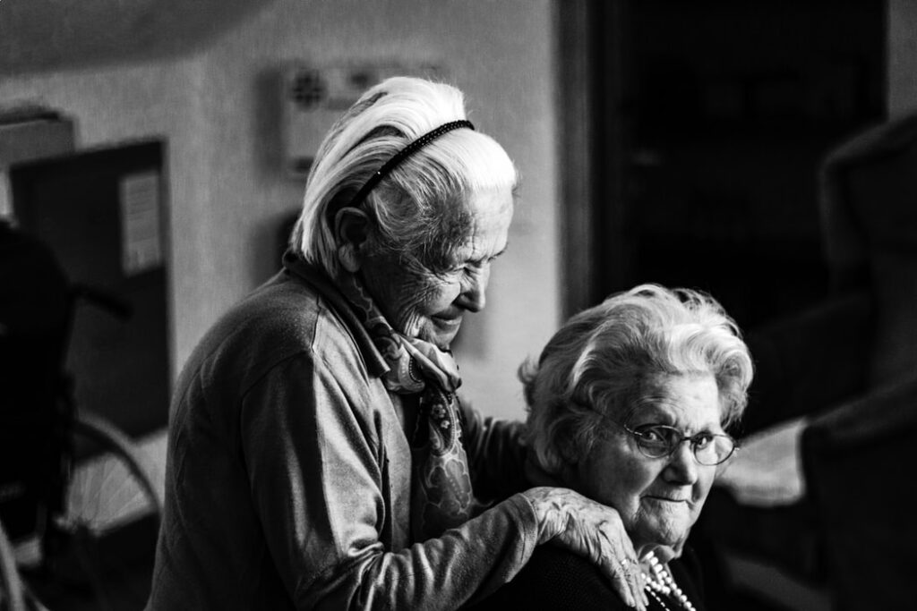 Duas senhoras idosas, uma está sentada e a outra em pé. A que está em pé apoia suas mãos nos ombros da que está sentada. Foto em preto e branco.