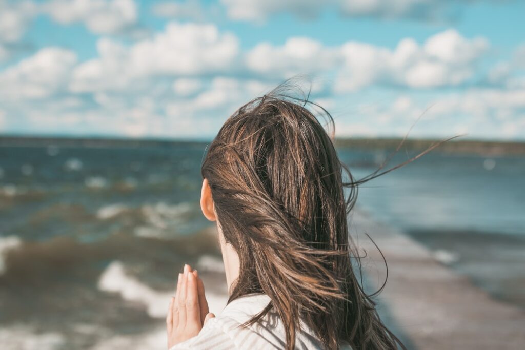 Mulher de costas olhando para o mar, e com as palmas das mãos unidas em frente ao seu corpo.