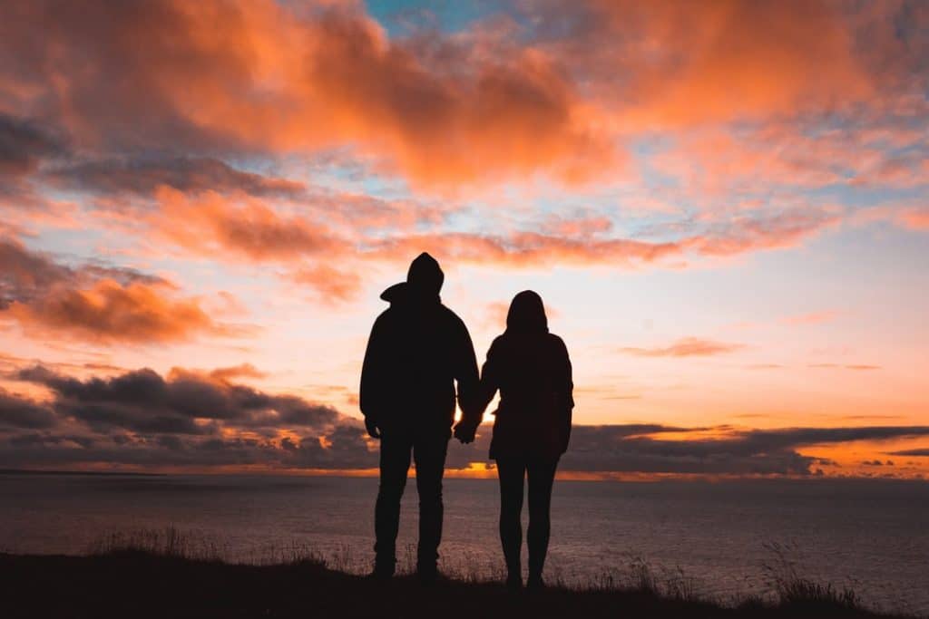 Silhueta de casal de mãos dadas de frente para o mar durante o pôr do sol.
