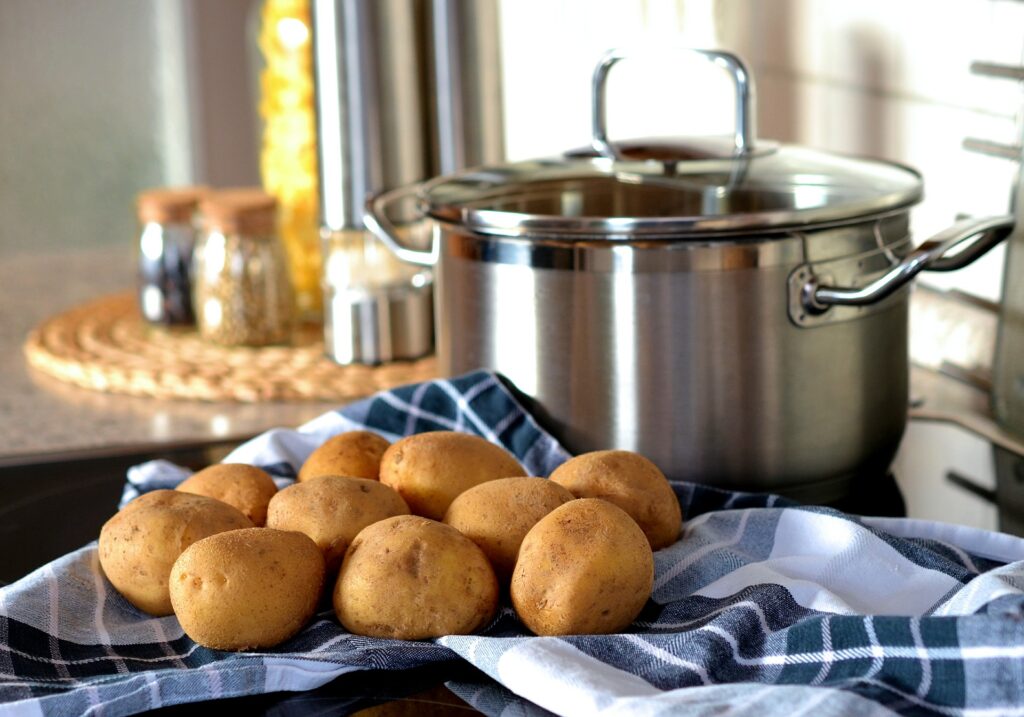 Batatas cruas sobre uma mesa forrada com uma toalha de pano nas cores azul e branco. Ao lado da porção de batatas uma panela grande de inox. Imagem de congerdesign por Pixabay.

