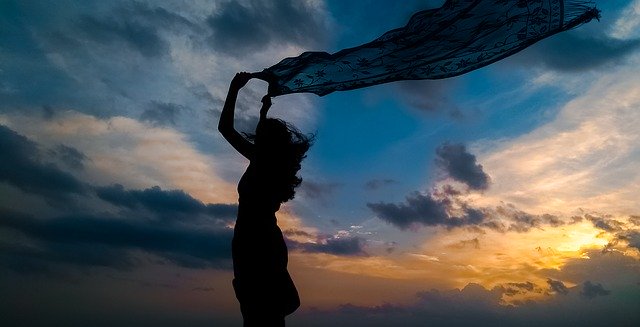 Silhueta de mulher com canga ao vento com céu com nuvens ao fundo 