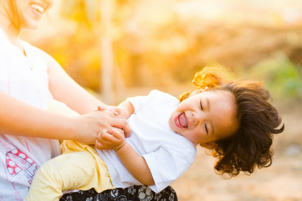 Criança brincando no colo da mãe, sorrindo e com os cabelos ao vente, em um parque ao pôr do sol.