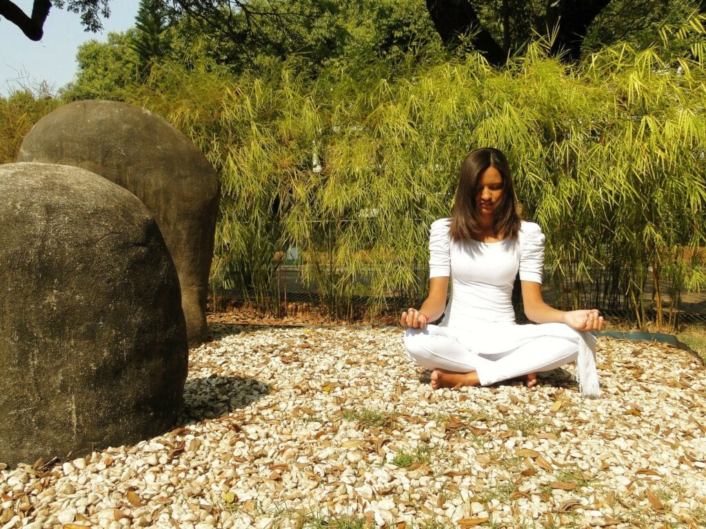Mulher vestida de branco sentada em posição de meditação. Ela está no meio da natureza. Ao fundo muitas árvores e ao lado pedras grandes.