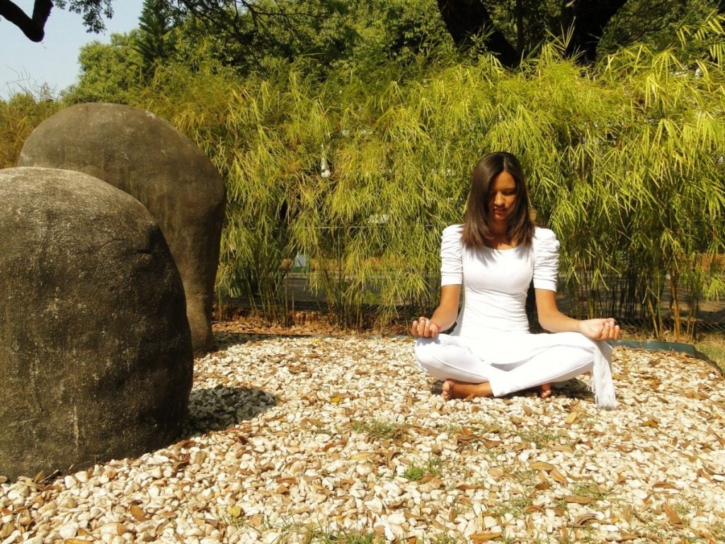 Mulher sentada com as pernas cruzadas em jardim com pequenas pedras brancas e folhas secas. Ela está meditando, e ao fundo muitos bambus plantados e árvores maiores.