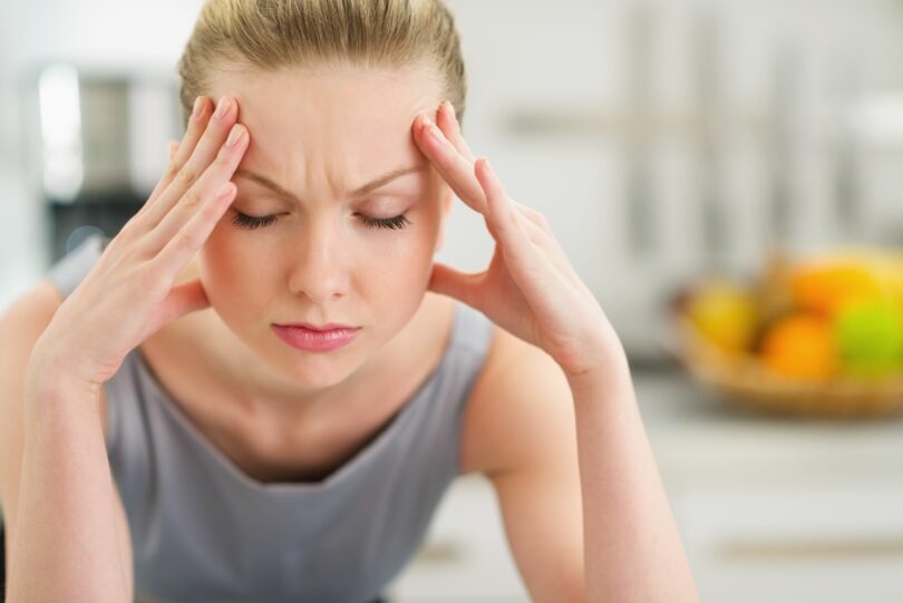 Mulher apoiada com seus cotovelos em uma bancada, olhos fechados e os dedos apoiados na testa.