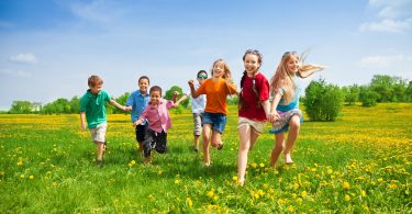 Grupo de crianças com quatro meninos e três meninas correndo em gramado durante o dia.