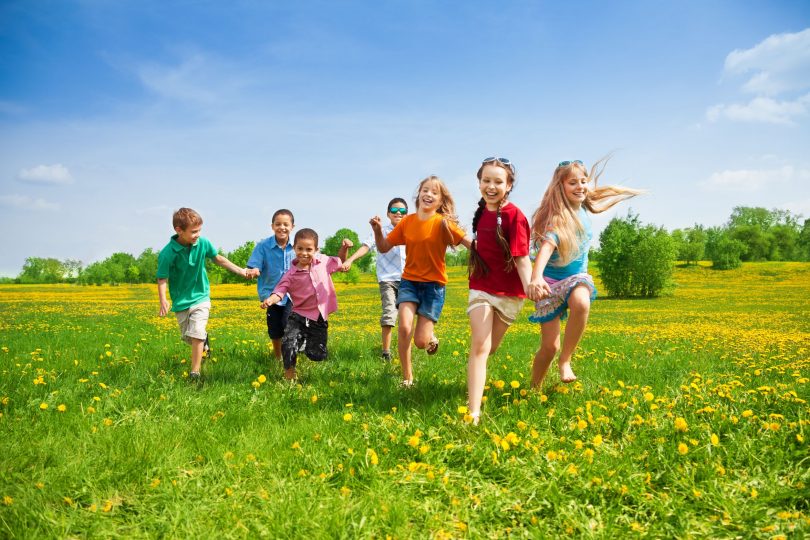 Grupo de crianças com quatro meninos e três meninas correndo em gramado durante o dia.