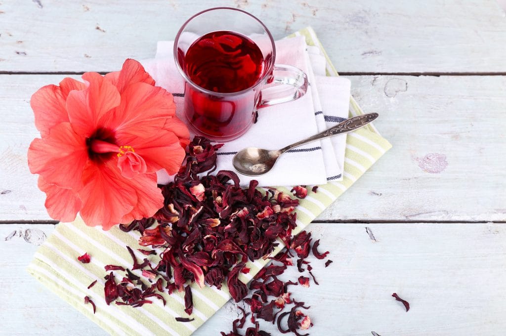 Chá de hibisco servido em uma caneca de vidro. Está sobre uma mesa de madeira pintada na cor brnaca. Ao lado da caneca, uma flor de hibisco e flores secas para o chá.