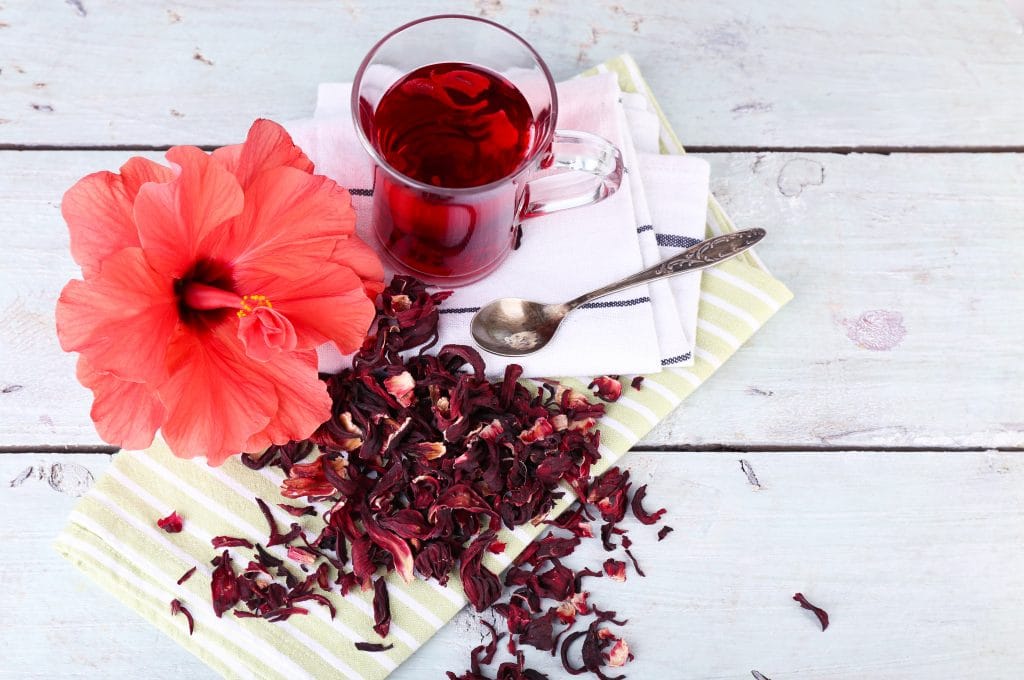 Chá de hibisco servido em uma caneca de vidro. Está sobre uma mesa de madeira pintada na cor brnaca. Ao lado da caneca, uma flor de hibisco e flores secas para o chá.
