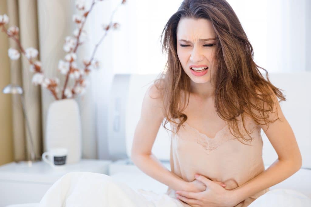Mulher dentro do seu quarto. Ela está sentada sobre a sua cama. Veste uma camisola na cor nude e aparenta está com muitas dores no estômago.