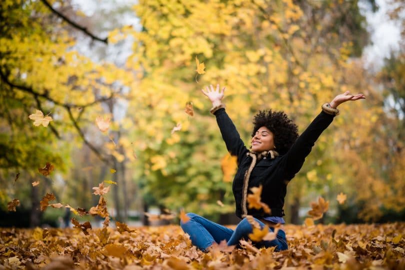 Mulher sentada no chão de parque com folhas de outono no chão e árvores ao fundo