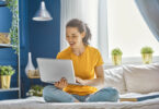 Mulher sentada com as pernas cruzadas em uma cama, segurando um notebook com sua mão direita, e digitando no mesmo com a mão esquerda.