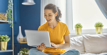 Mulher sentada com as pernas cruzadas em uma cama, segurando um notebook com sua mão direita, e digitando no mesmo com a mão esquerda.