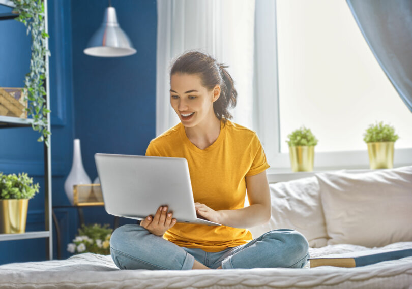 Mulher sentada com as pernas cruzadas em uma cama, segurando um notebook com sua mão direita, e digitando no mesmo com a mão esquerda.