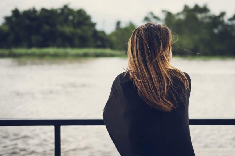 Mulher de costas, apoiada em uma barreira de metal, olhando para um rio na sua frente.